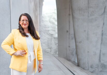 Frau mit schwarzen Haaren lächelt in die Kamera. Sie trägt eine Brille, einen gelben Blazer und weißen Top
